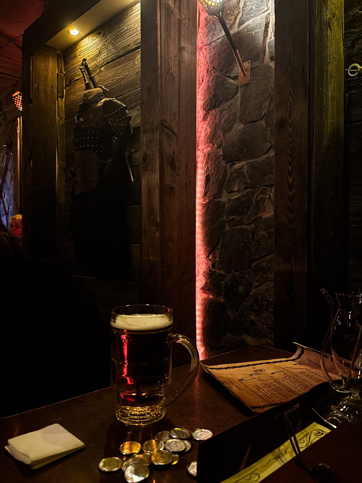 Chope de bière posée sur une table en bois au milieu de l'ambiance médiévale de la Tawerna Wilczy Dół, avec armure exposée et mur de pierre éclairé de rouge