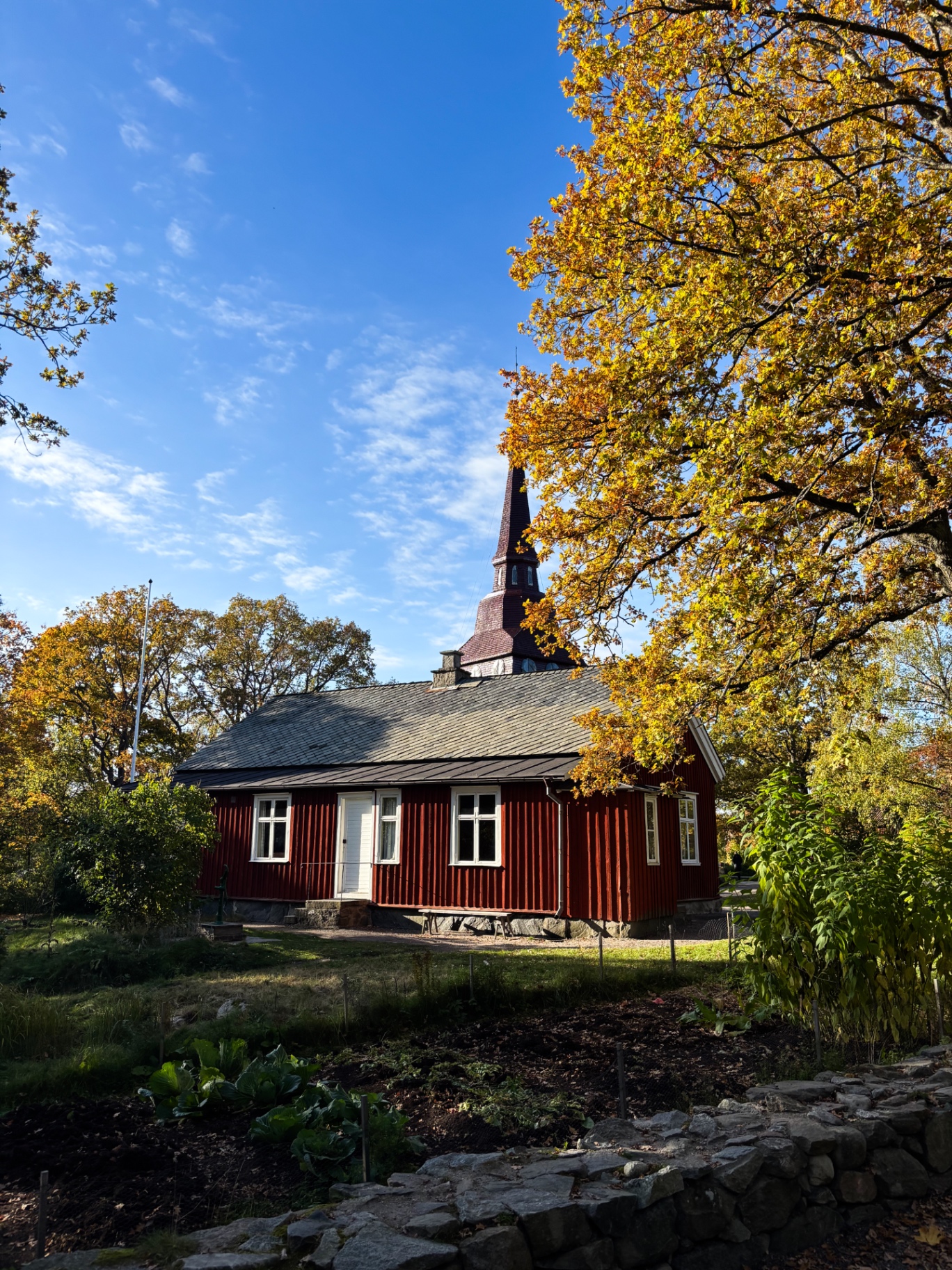 Maison rouge en bois de Skansen, au pied d’un clocher, sous les arbres dorés