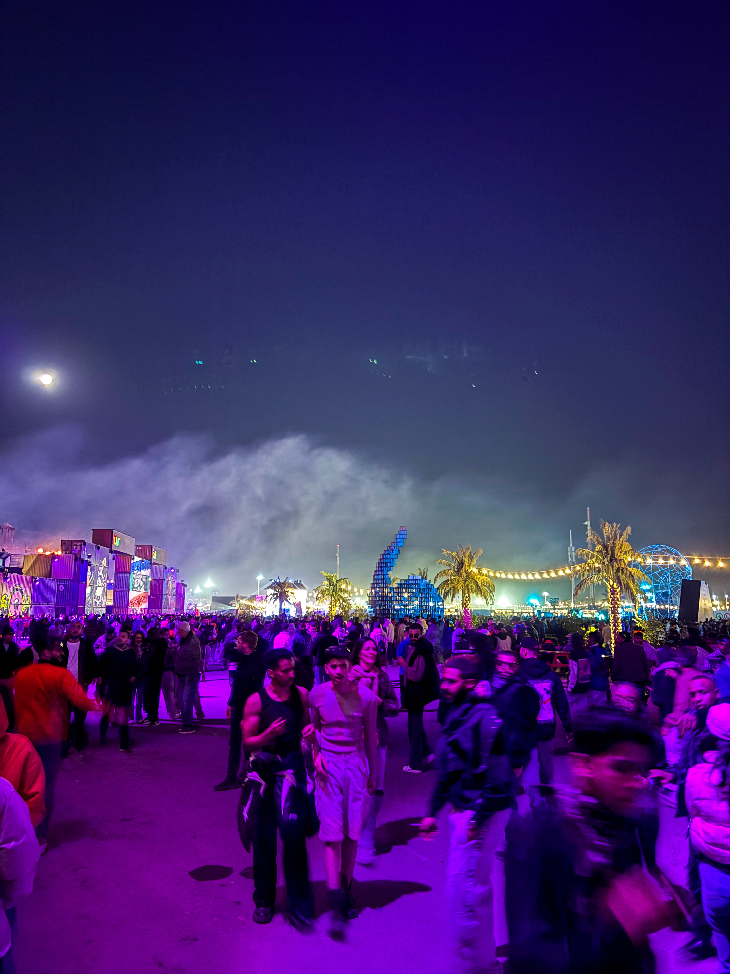 Festivaliers dans les allées de Soundstorm sous la lune, entre installations lumineuses et palmiers
