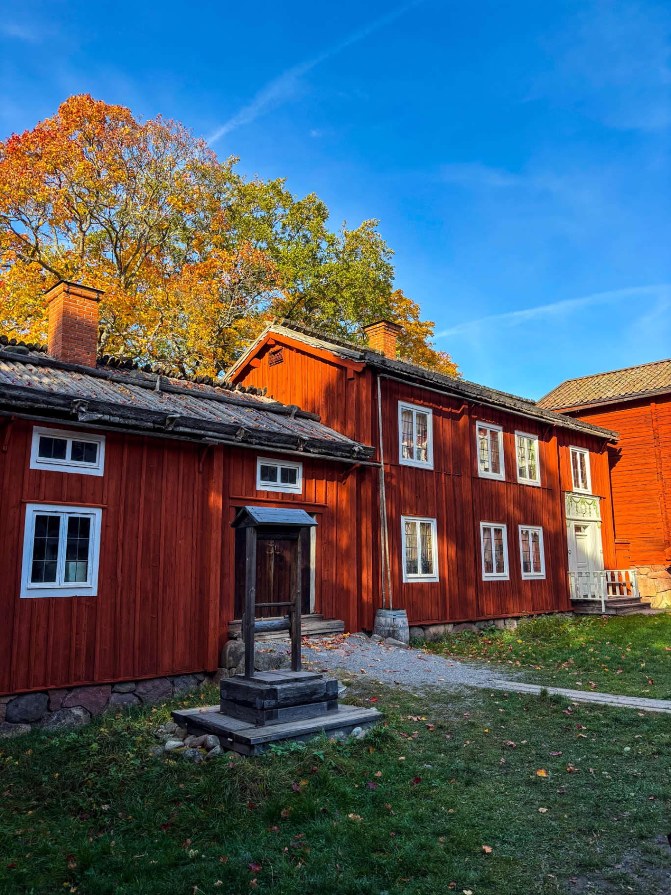Grande demeure rouge à pans de bois avec puits en bois et arbres d’automne, à Skansen