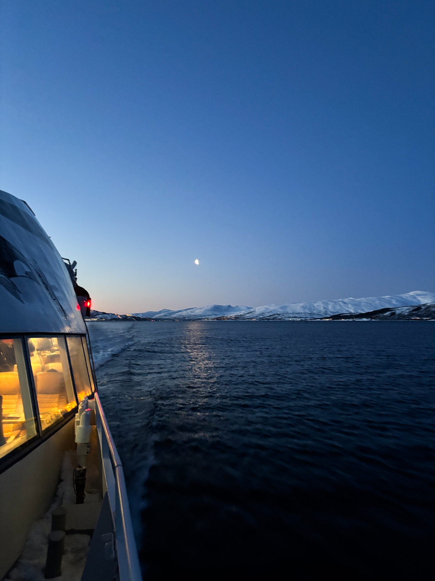 Bateau naviguant au crépuscule vers les fjords du nord, lune et montagnes enneigées à l’horizon