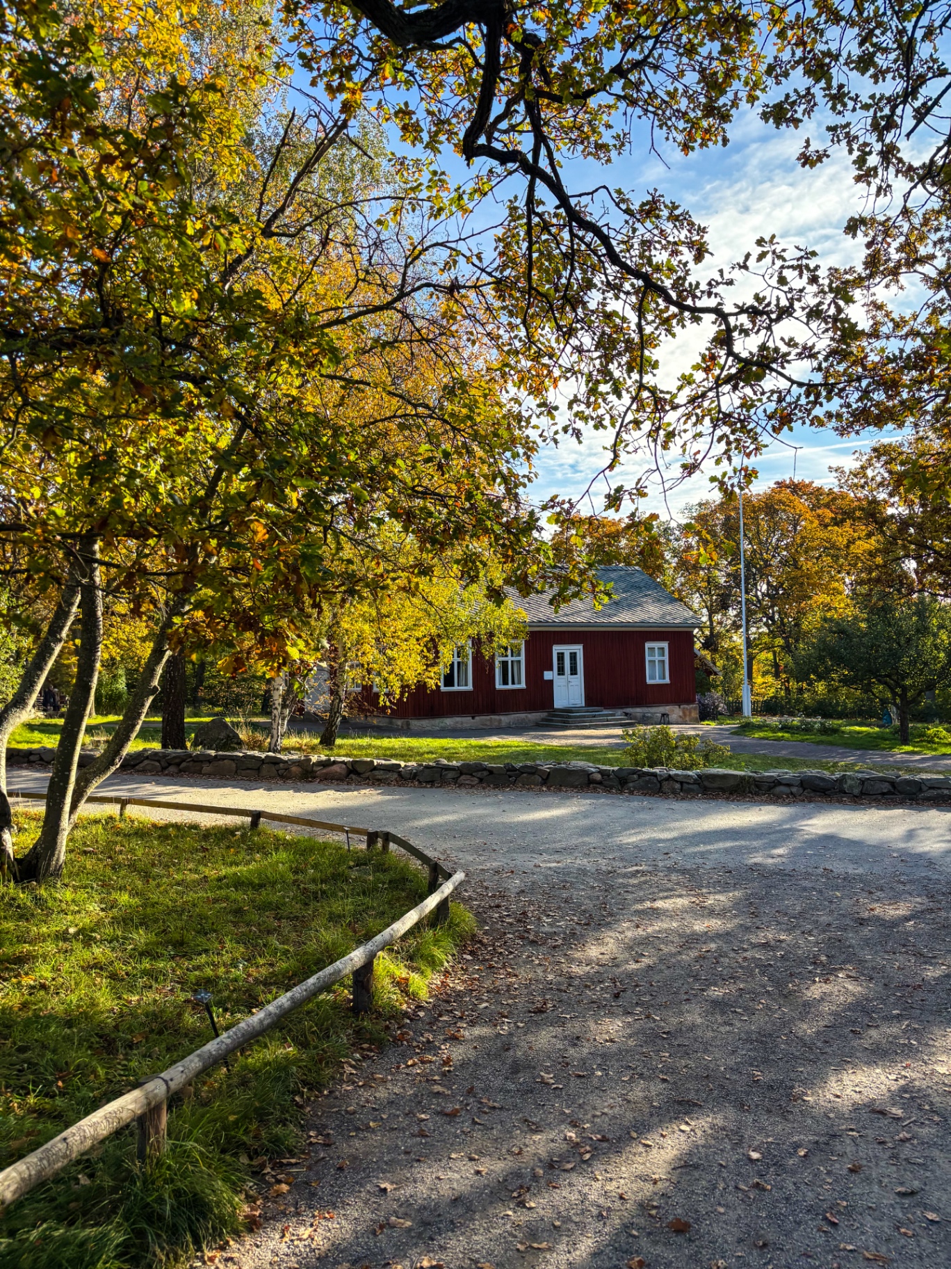 Sentier bordé de murets de pierre menant à une maison rouge sous les feuilles dorées, à Skansen