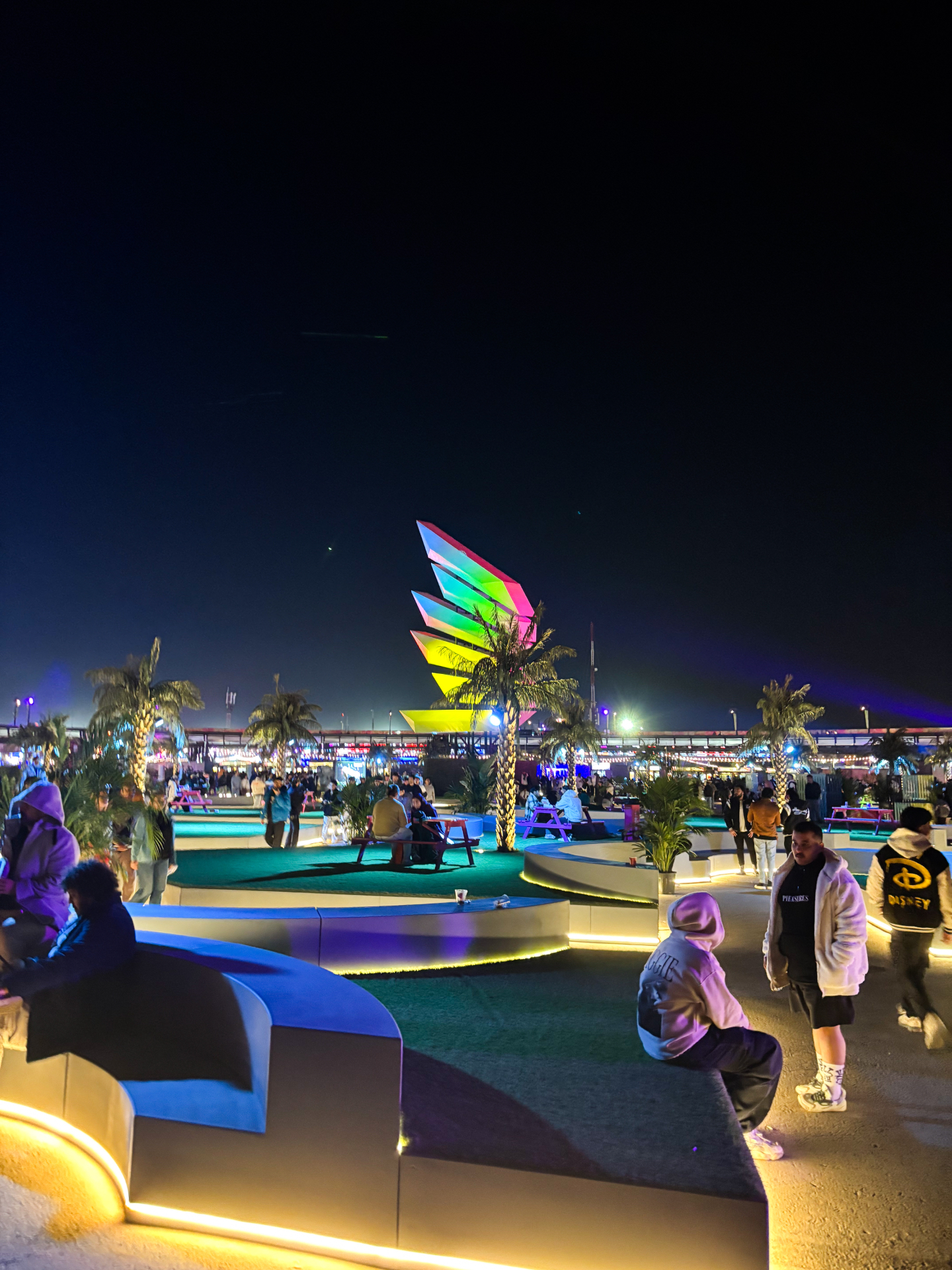 La grande place de Soundstorm la nuit, avec bancs lumineux, tables de pique-nique et sculpture multicolore en forme d'ailes